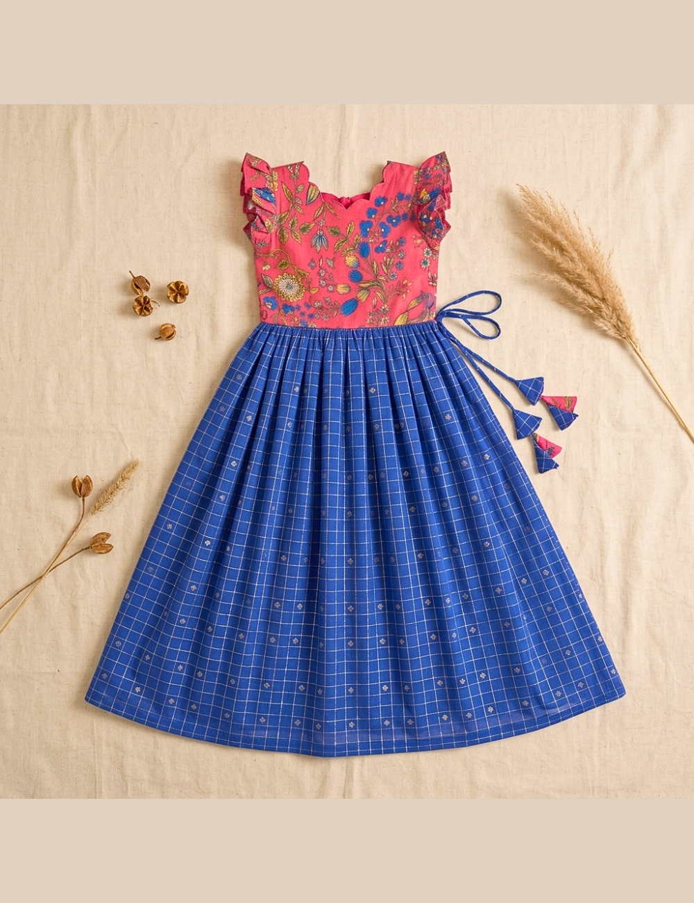 A girls' sleeveless dress laid flat on a beige fabric background with dried decorative grass and small flowers. The dress has a bright pink floral bodice with ruffled shoulders and a blue plaid skirt with small white motifs and blue tassel ties at the side.
