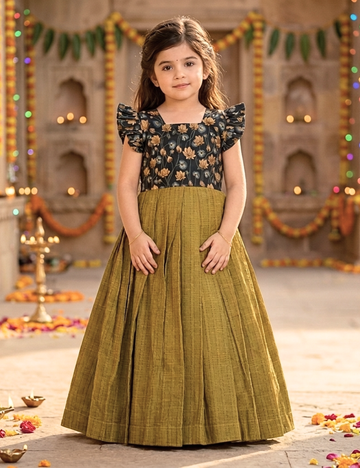 A young girl modeling a traditional Indian dress with a black floral bodice and a pleated olive green skirt, standing in a decorated courtyard.