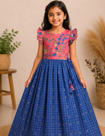 A young girl modeling a traditional Indian dress with a pink floral embroidered bodice and a blue checkered flared skirt, set against a warm beige background with decorative plants.