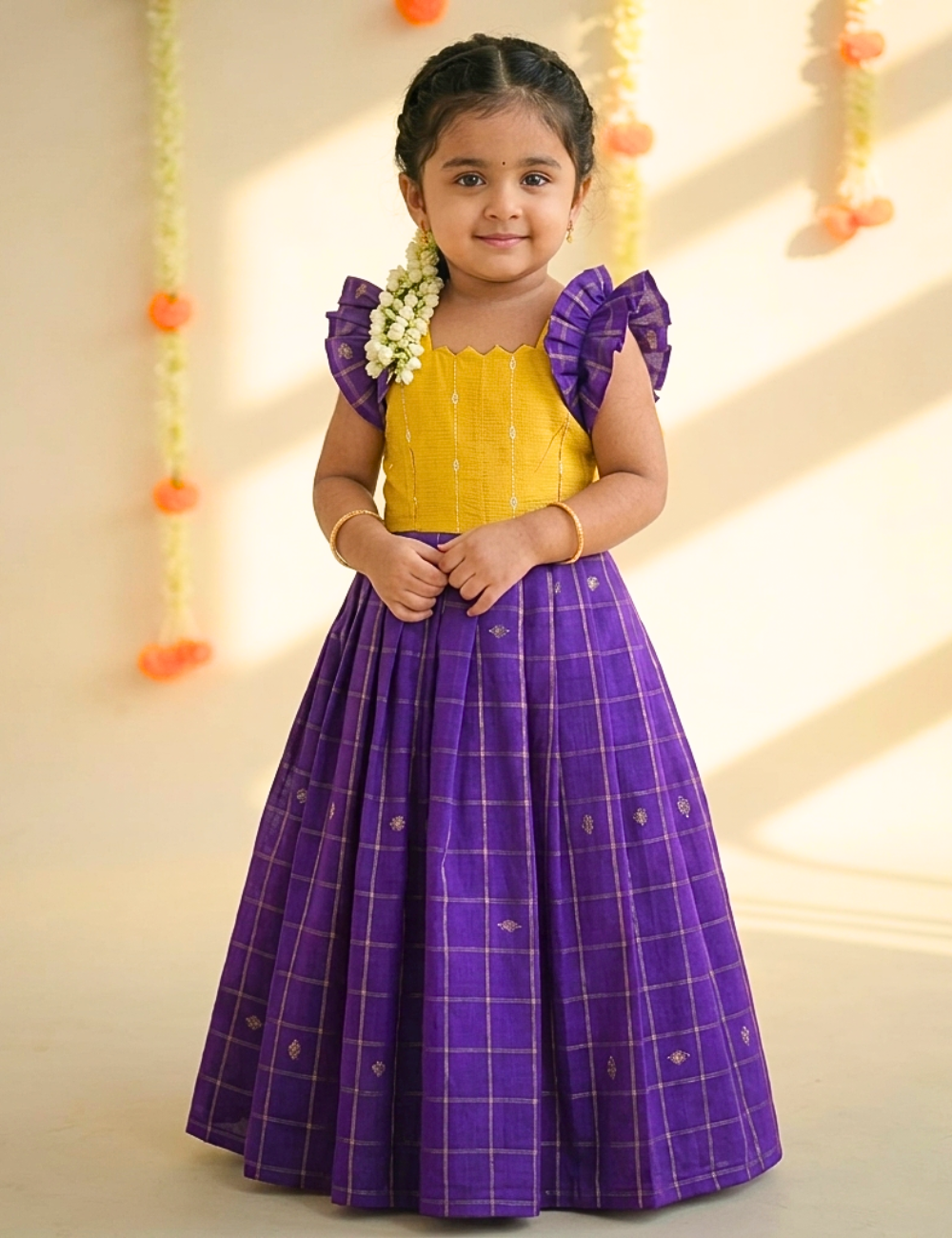 A young girl modeling a traditional Indian outfit featuring a bright yellow sleeveless top with purple ruffled shoulder details and a voluminous purple skirt with a subtle gold grid pattern, set against a warm beige background with hanging marigold garlands.