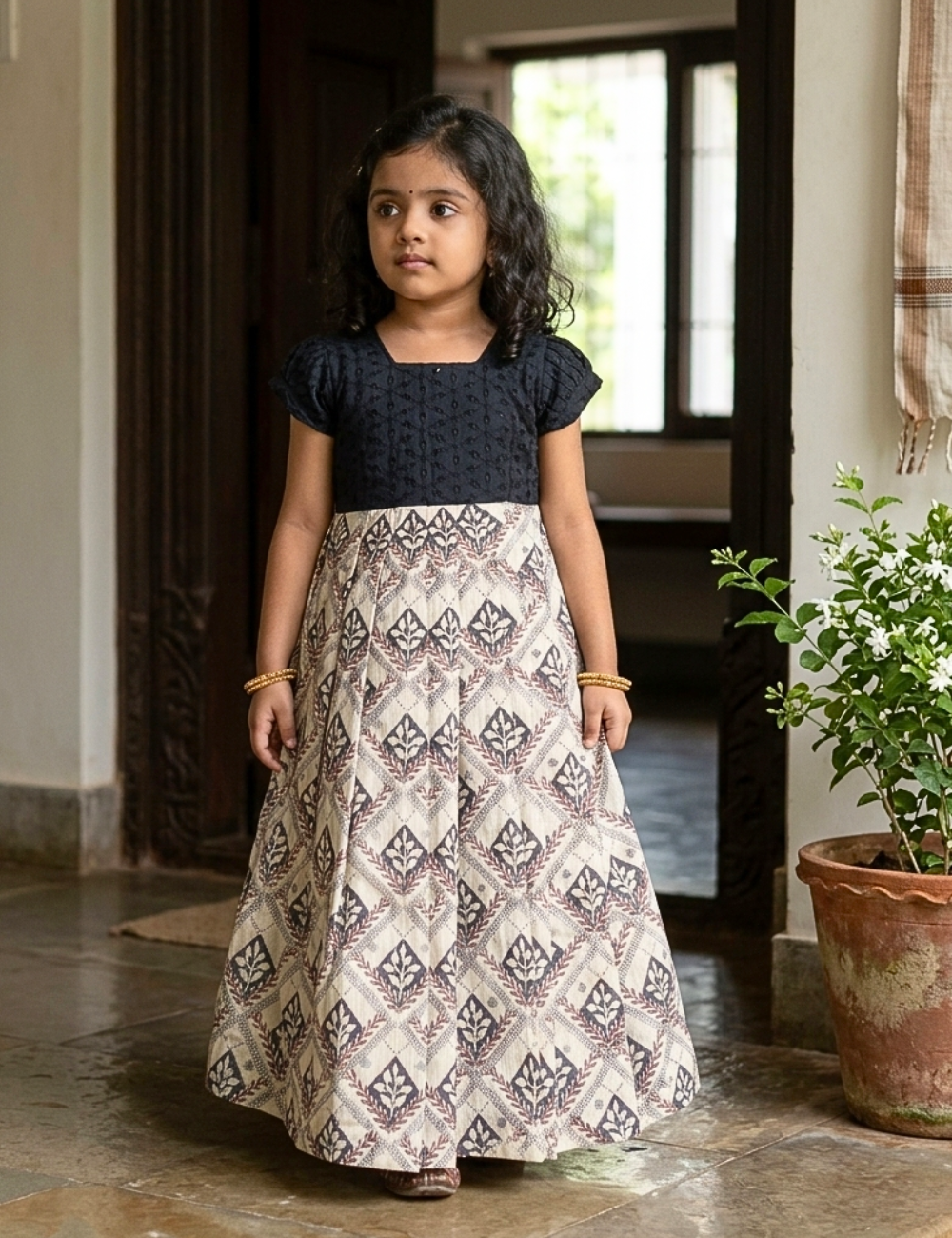 A young girl stands smiling in a decorated indoor setting, wearing a traditional Indian dress with a dark navy blue textured short-sleeve top and a voluminous skirt featuring a beige, gray, and black geometric ikat pattern. The background includes hanging brass bells, marigold garlands, and decorative bowls.