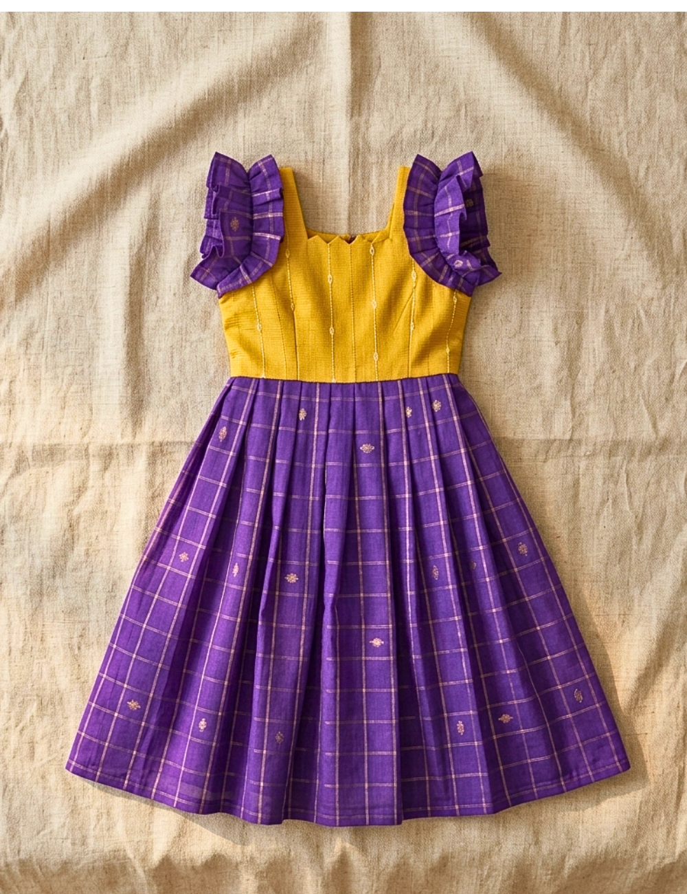 A sleeveless children's dress laid flat on a beige linen background. The bodice is mustard yellow with ruffled shoulder details, and the full skirt is purple with a thin gold grid pattern and small gold motifs.