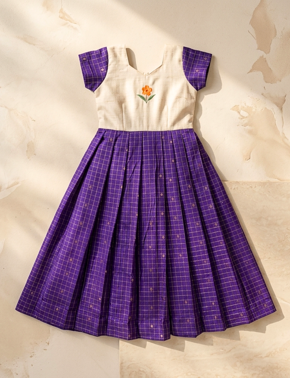 A vintage-style children's dress with a cream-colored bodice featuring an embroidered orange flower, and a pleated purple skirt with a white grid pattern. The dress is laid flat on a textured beige background with soft shadows.