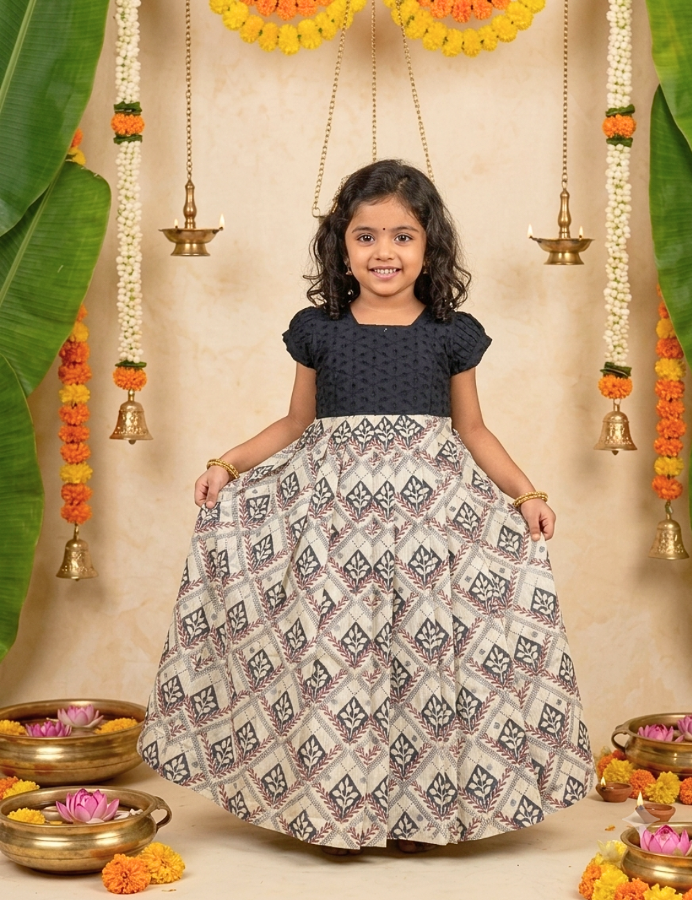 A young girl stands smiling in a decorated indoor setting, wearing a traditional Indian dress with a dark navy blue textured short-sleeve top and a voluminous skirt featuring a beige, gray, and black geometric ikat pattern. The background includes hanging brass bells, marigold garlands, and decorative bowls.