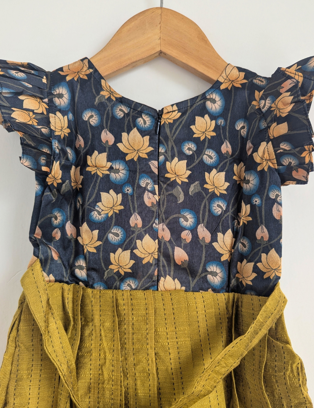 A children's dress hanging on a wooden hanger against a plain white wall. The top portion has a dark navy blue base with a floral print in beige, blue, and pink, while the lower skirt portion is a textured mustard yellow fabric.