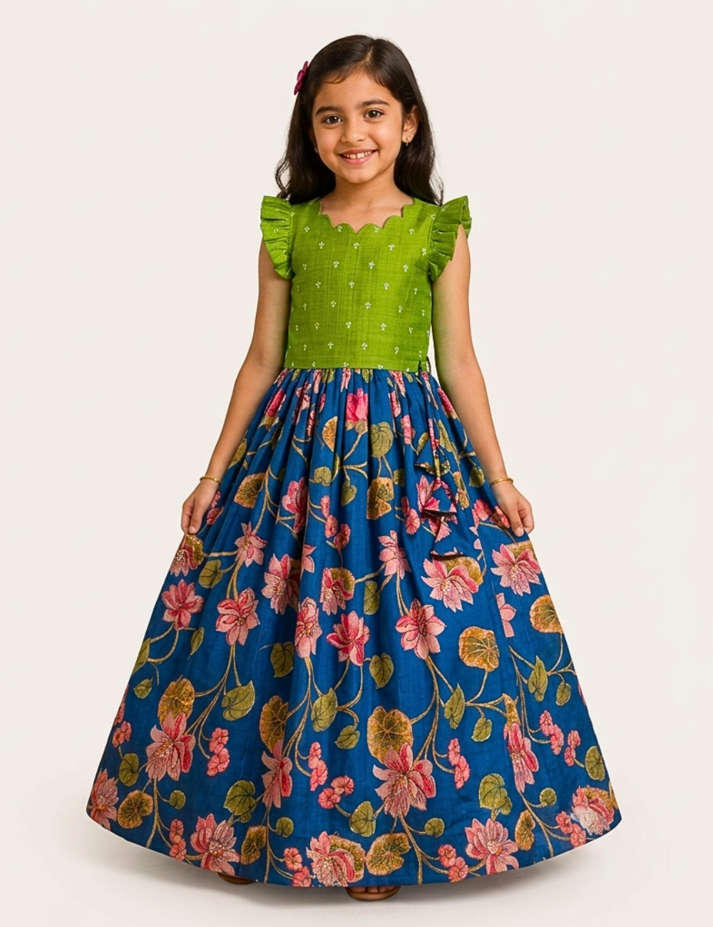 A young girl modeling a long dress with a bright green ruffled bodice and a full blue skirt featuring a pink and green floral print, set against a plain light gray background.