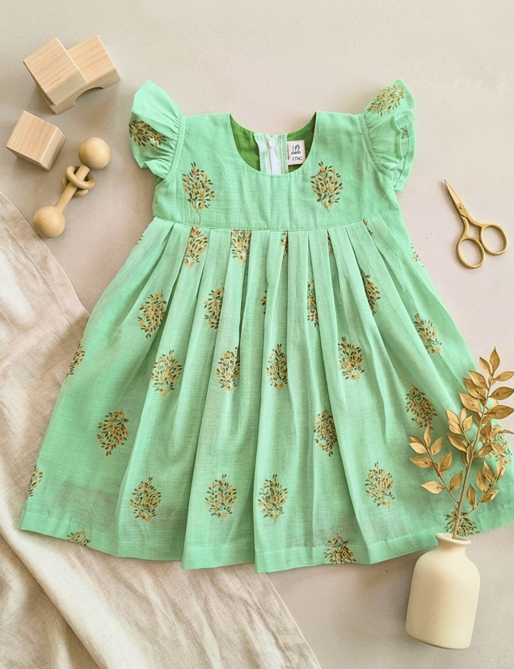 A mint green baby dress with gold floral prints, laid flat on a beige surface with styled props including wooden blocks, scissors, and a small vase with dried leaves.