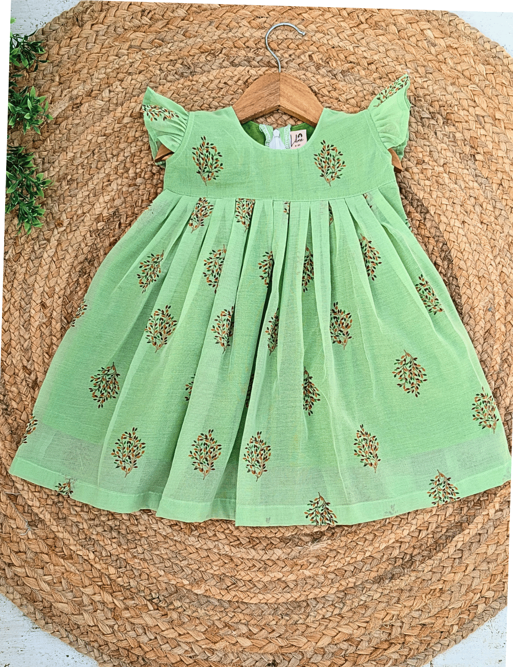 A light green cotton girl's dress with gold floral embroidery, displayed on a wooden hanger over a round woven straw mat.
