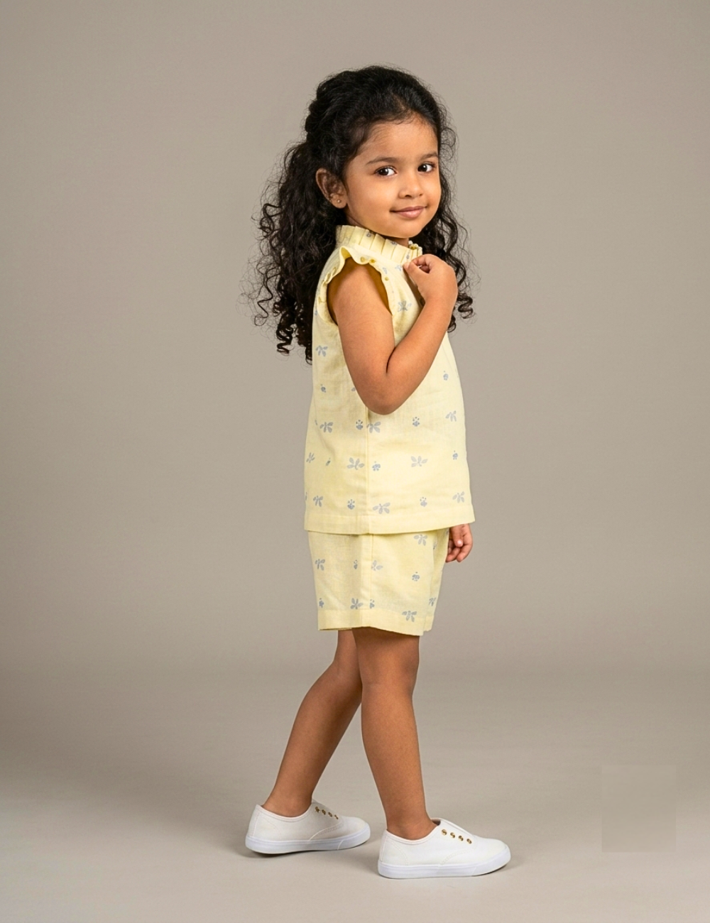 A young girl with curly dark hair stands in profile wearing a pale yellow sleeveless top and matching shorts with a small blue floral pattern, paired with white sneakers, against a plain beige studio background.