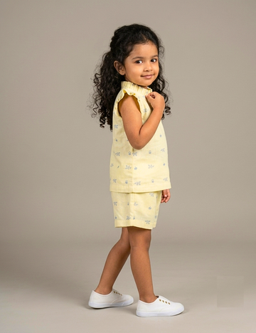 A young girl with curly dark hair stands in profile wearing a pale yellow sleeveless top and matching shorts with a small blue floral pattern, paired with white sneakers, against a plain beige studio background.