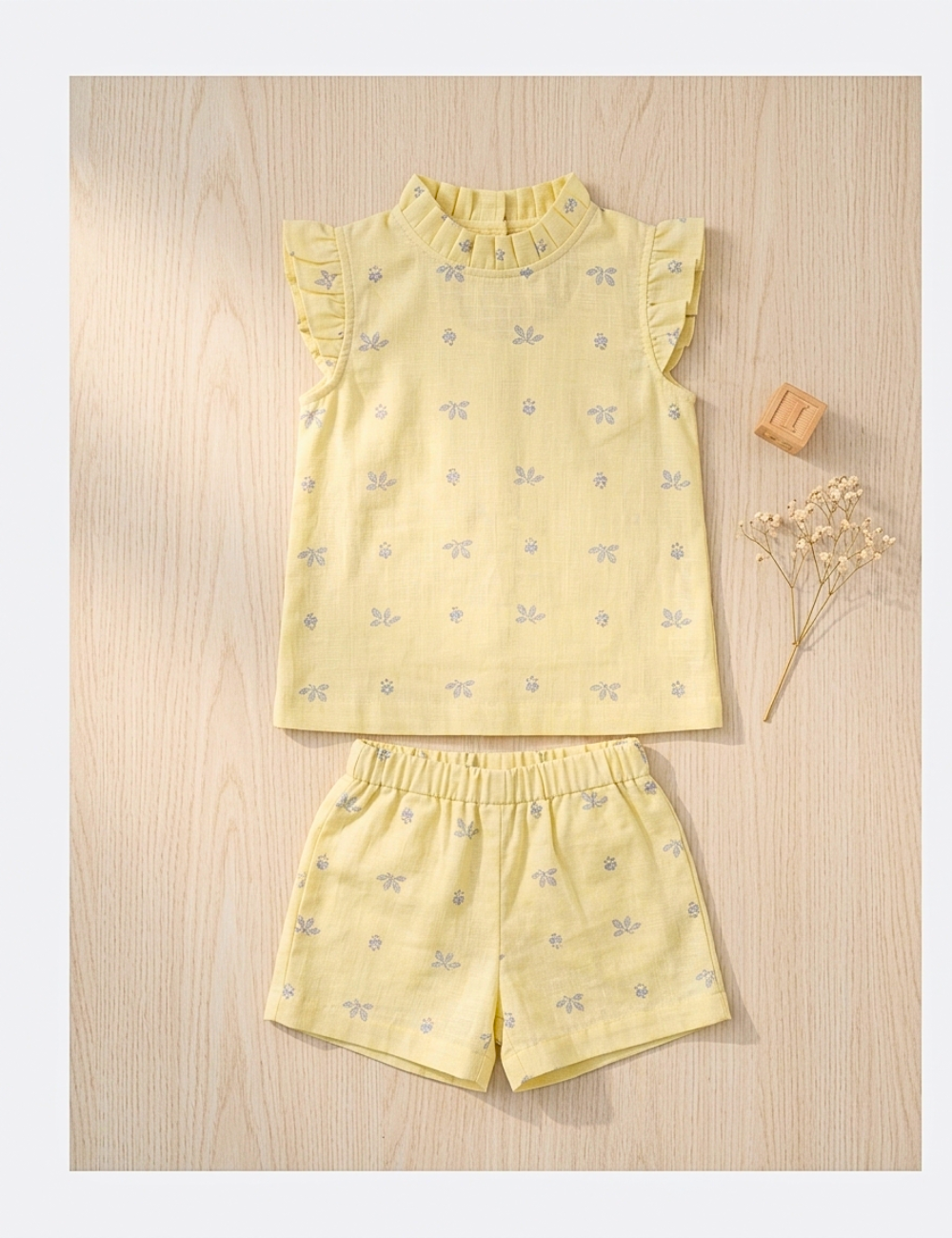 A two-piece baby girl's outfit set consisting of a pale yellow sleeveless top with ruffled shoulders and matching shorts, both decorated with a small light blue bow pattern, laid flat on a light wood background.