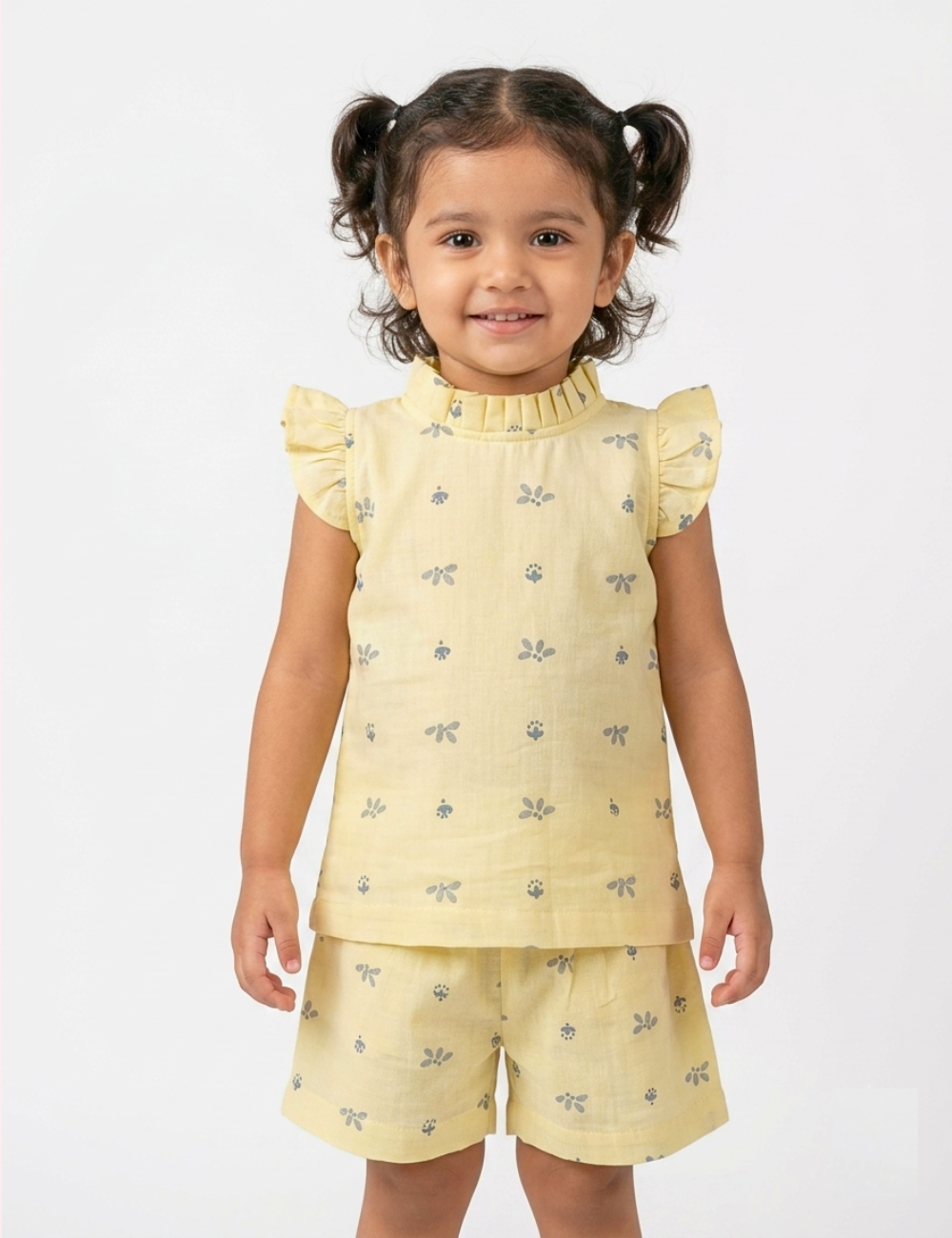 A young girl with dark hair in pigtails wearing a light yellow two-piece set with a small gray dragonfly pattern, consisting of a sleeveless top with ruffled shoulders and matching shorts, against a plain white background.