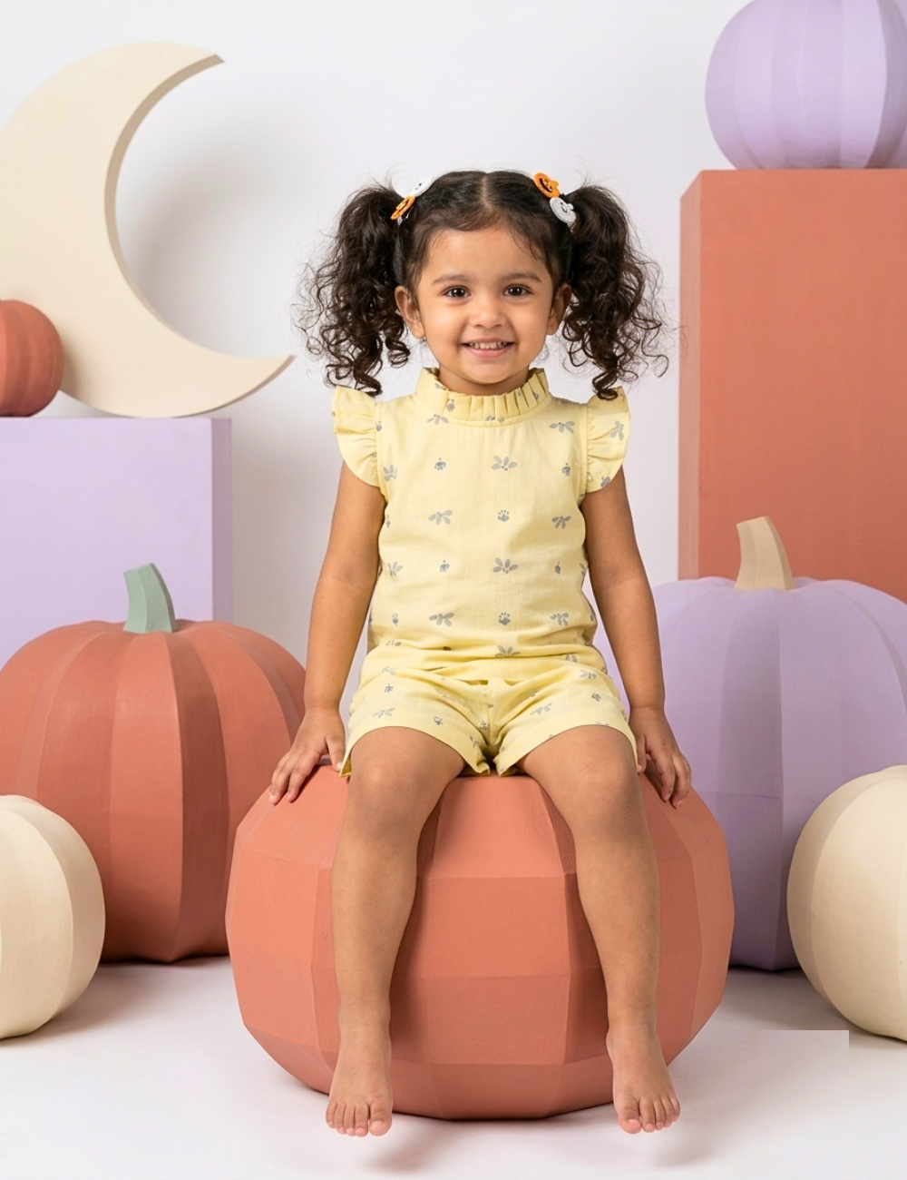 A young girl with curly dark hair in pigtails sits on a peach pumpkin-shaped bean bag chair. She is wearing a pale yellow short-sleeve romper with a small butterfly pattern.
