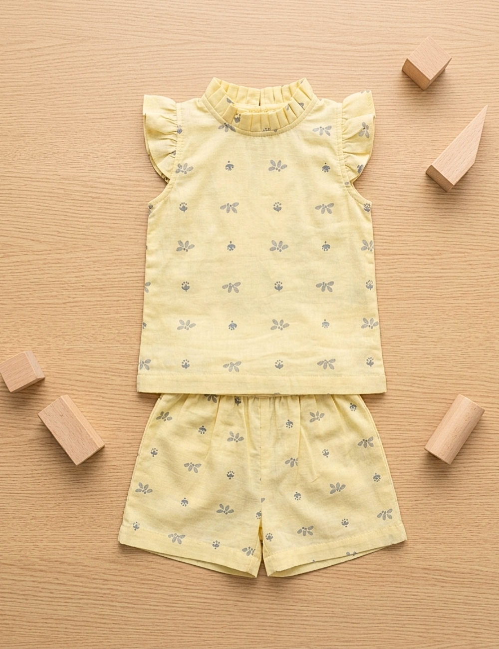 A baby girl's two-piece clothing set consisting of a sleeveless top with ruffled shoulders and matching shorts, both in pale yellow with a small gray bow pattern, laid flat on a light wood background with wooden toy blocks.