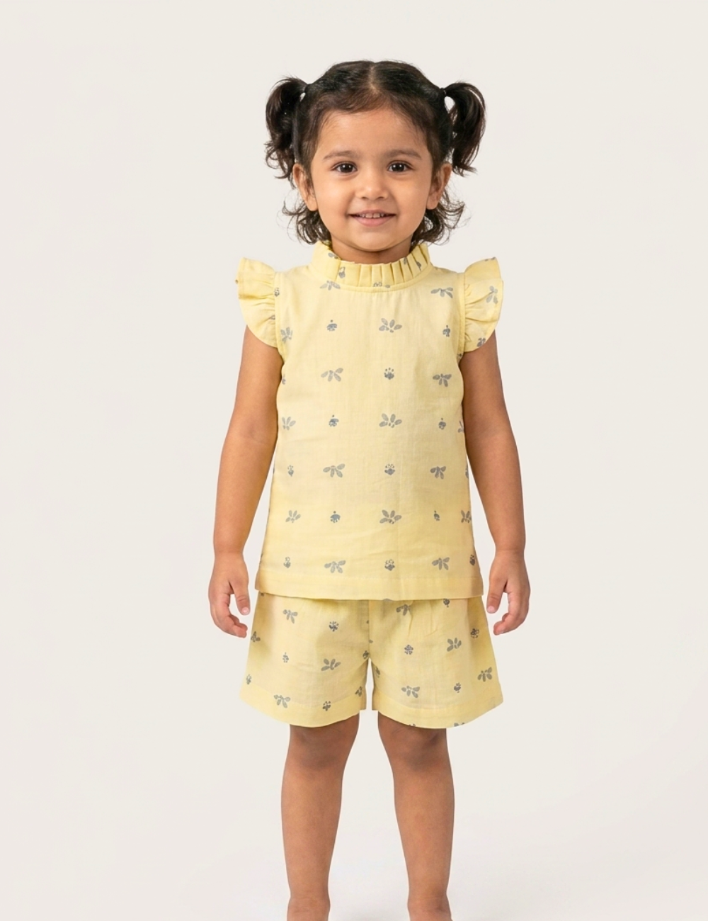 A young girl with dark curly hair in pigtails stands against a light gray studio background wearing a pale yellow two-piece outfit with a small blue pattern. The set includes a short-sleeve top with ruffled shoulders and matching shorts.